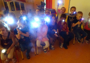 Grupa dzieci siedzi na krzesłach, w ręku trzymają włączone latarki.